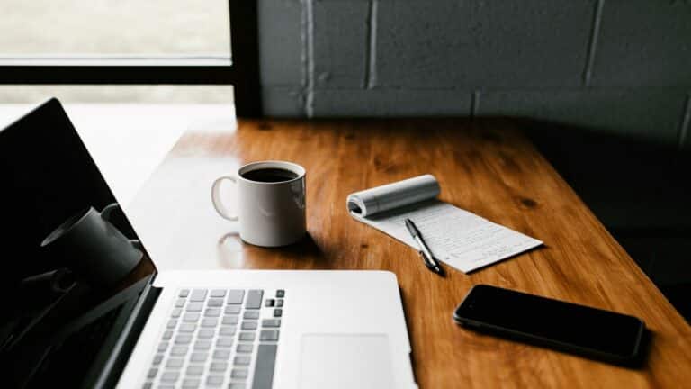 A workspace with a laptop, coffee mug, notepad, pen, and smartphone on a wooden table near a window.