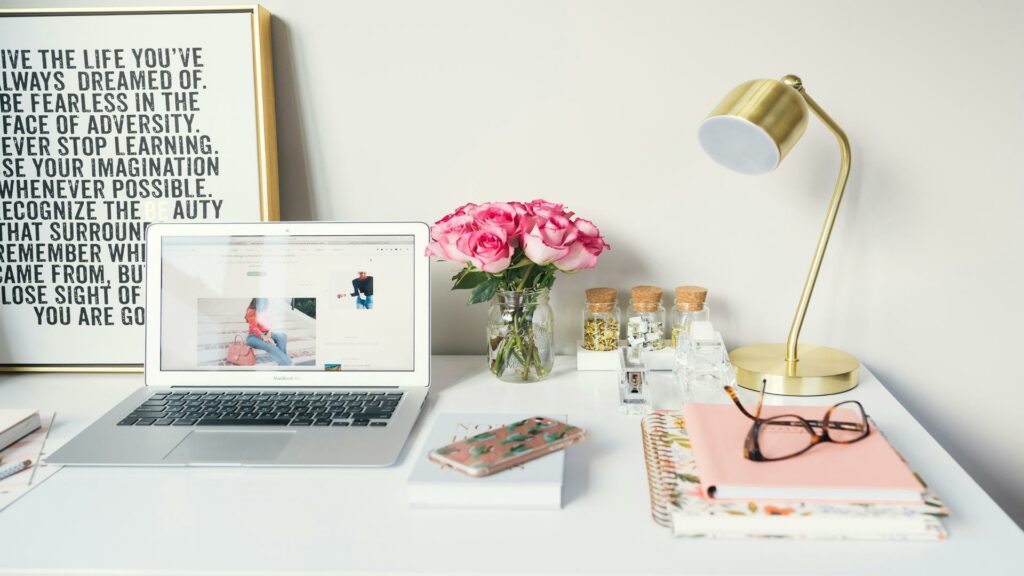 A clean workspace with a laptop, pink roses, a gold lamp, and notebooks.