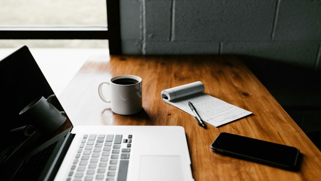 A wooden desk with a laptop, coffee mug, notepad, pen, and smartphone near a window.