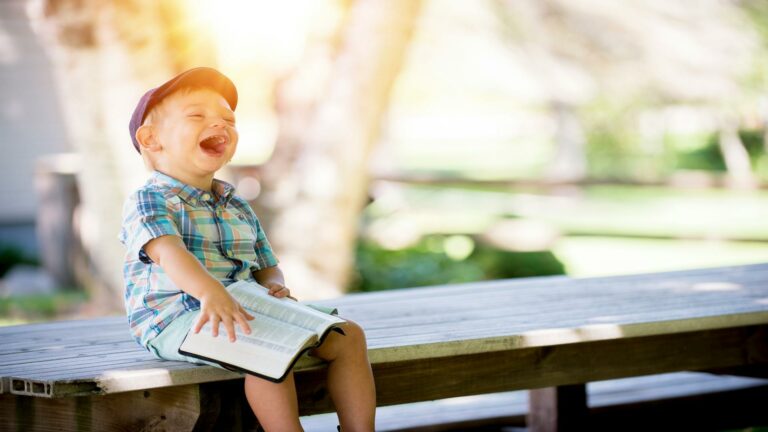 Young child laughing while reading a book outdoors on wooden bench