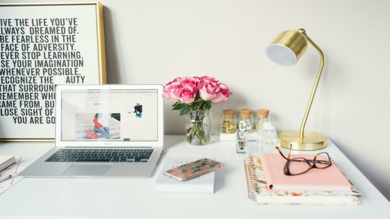 A stylish desk setup with a laptop displaying online customer reviews, pink roses, and a gold lamp.