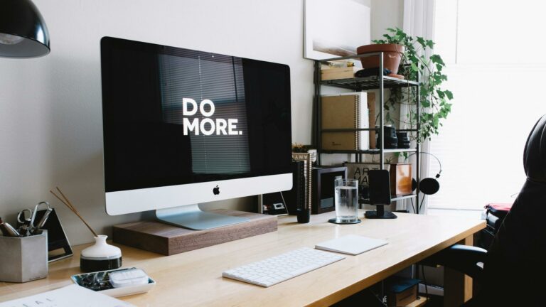 Modern workspace setup with iMac displaying 'DO MORE' text on screen, featuring wooden desk, plants, and office accessories