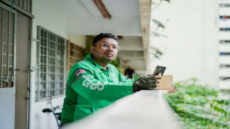 A delivery driver in a green uniform holding a phone and package