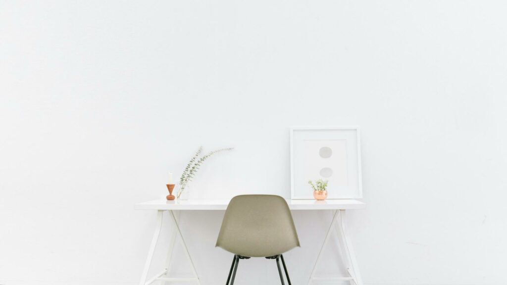 A minimalist desk setup with a chair, small plants, and a framed artwork on a white background.