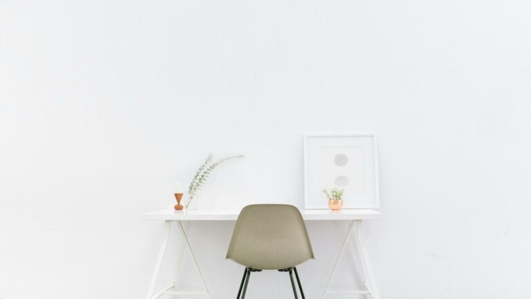 A minimalist desk setup with a chair, small plants, and a framed artwork on a white background.