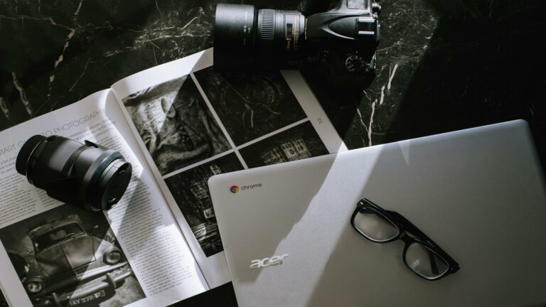 Flatlay of photography equipment including camera lenses, Acer Chromebook, photography magazine, and black-rimmed glasses on dark marble surface