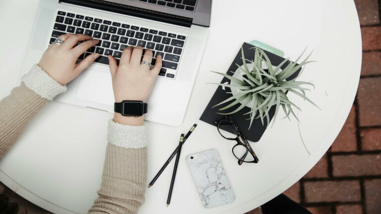 Person typing on a laptop with tools for content creation services on a white desk