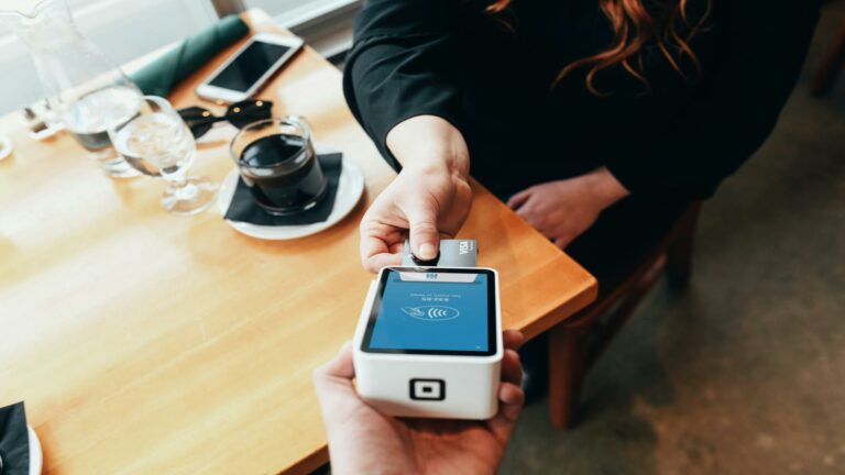 Person making a payment using a card reader at a table with drinks and a smartphone nearby.