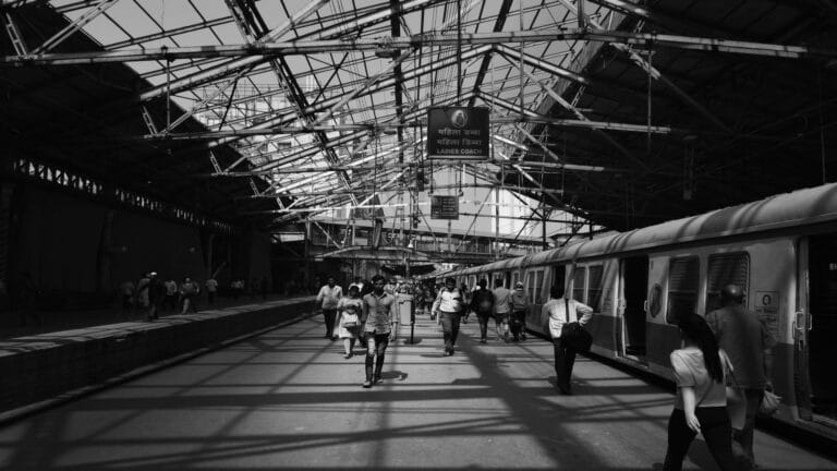 Black and white photograph of a railway platform with Ladies Coach sign in Hindi and English, showing passengers walking along a train platform under a glass roof structure