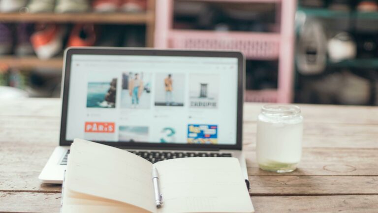 Laptop displaying visual content grid with notebook and glass jar on wooden desk