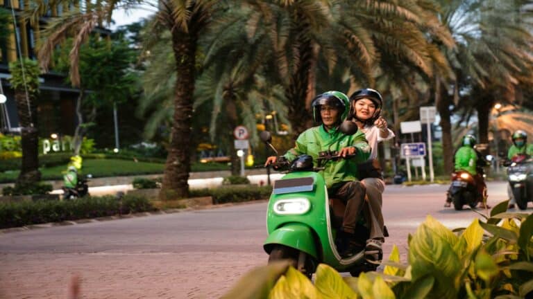 Two people riding a green scooter on a city street lined with palm trees