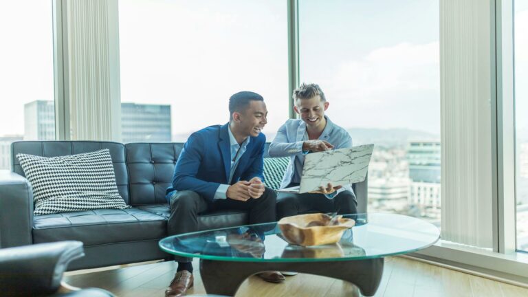 Two young professionals in business attire discussing website security on a laptop in a modern office setting with city views