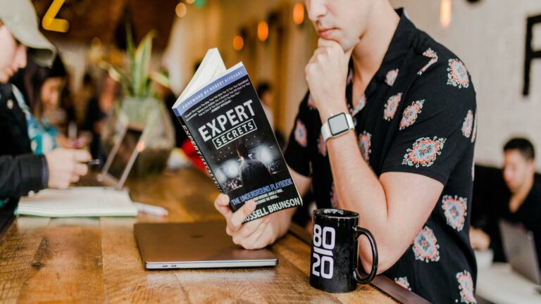 Person in patterned shirt reading Expert Secrets book with laptop and 80/20 coffee mug on wooden table