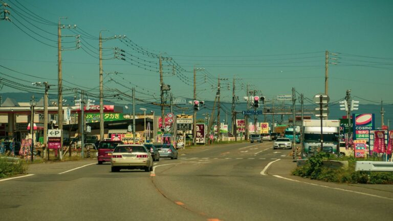 Wide suburban road in Japan with multiple power lines, Japanese language signs, retail stores, and cars on a clear day with teal-colored sky