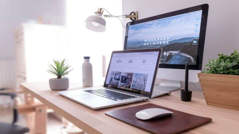 Website design agency professional desk setup with MacBook, monitor displaying 'Work Hard Anywhere', desk lamp, and minimalist decor