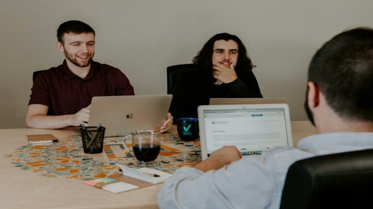 Three content marketing professionals working together at a table with laptops and coffee, discussing strategy in a modern office setting