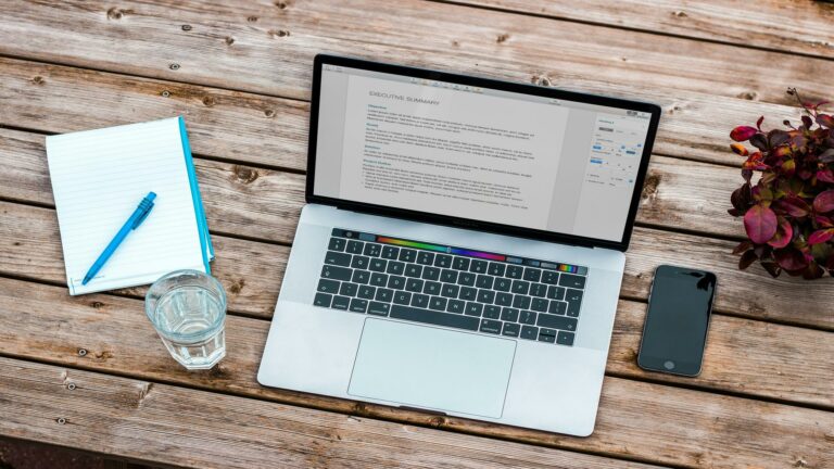 Laptop on wooden table with notebook, pen, smartphone, and plant