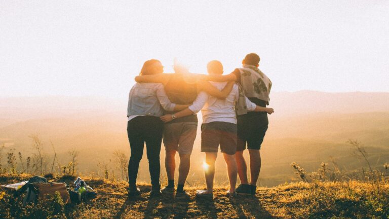 A group of five people standing together with arms around each other, facing a sunset.