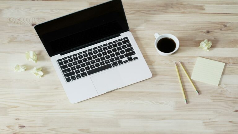 A laptop on a wooden desk with crumpled paper, pencils, a notepad, and a coffee cup.