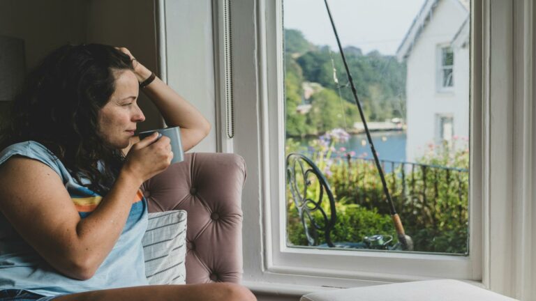 Woman sipping coffee while sitting by a window overlooking a garden and a lake.