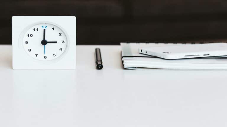 Minimalist desk setup with white analog clock showing 3:15, black pen, and mobile device on white surface