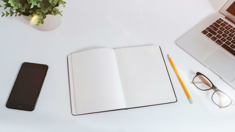 Minimalist desk setup with open blank notebook, smartphone, laptop corner, reading glasses, and small plant