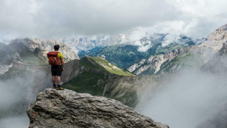 A hiker with a red backpack standing on a rocky cliff, overlooking a misty mountain range.