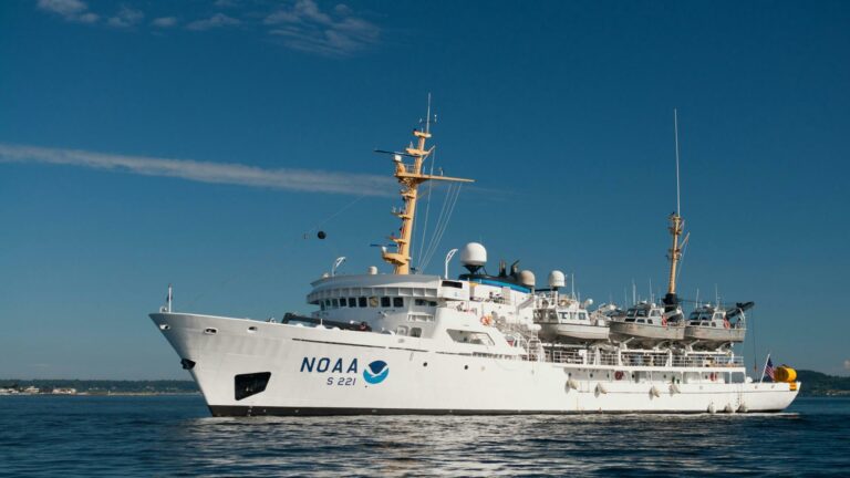 NOAA research vessel with a white hull and yellow mast sailing on calm waters under a clear blue sky.