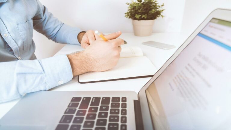 Person writing notes with a laptop open on a desk