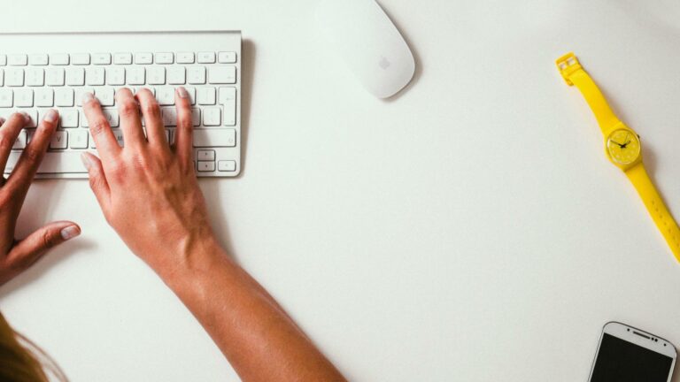 Person typing on white keyboard with wireless mouse and yellow watch on white desk