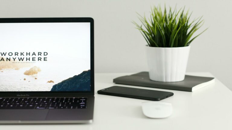 Minimalist workspace setup with MacBook Pro displaying 'Work Hard Anywhere' text, smartphone, potted plant, and wireless mouse on white desk