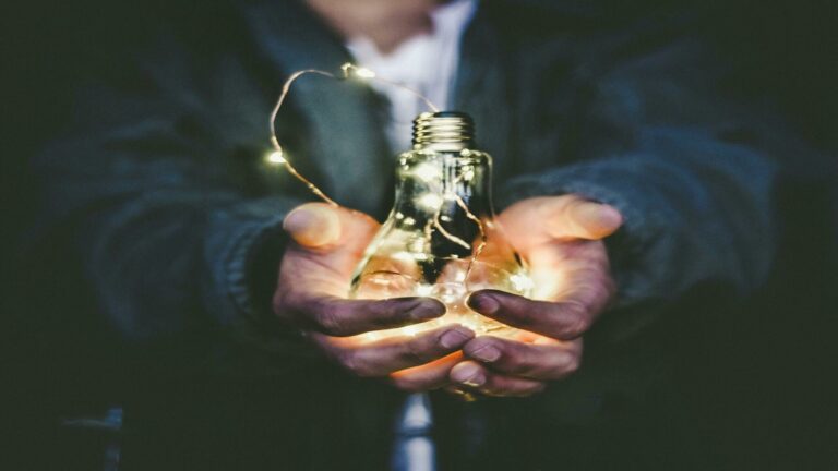 Hands holding illuminated light bulb with fairy lights inside against dark background