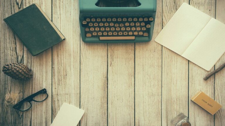 A flat-lay of a vintage typewriter, notebook, book, glasses, pinecone, and other items on a wooden table