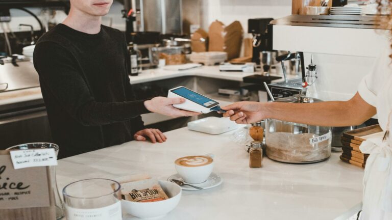 A person handing a payment card to a barista using a card reader in a cafe setting.