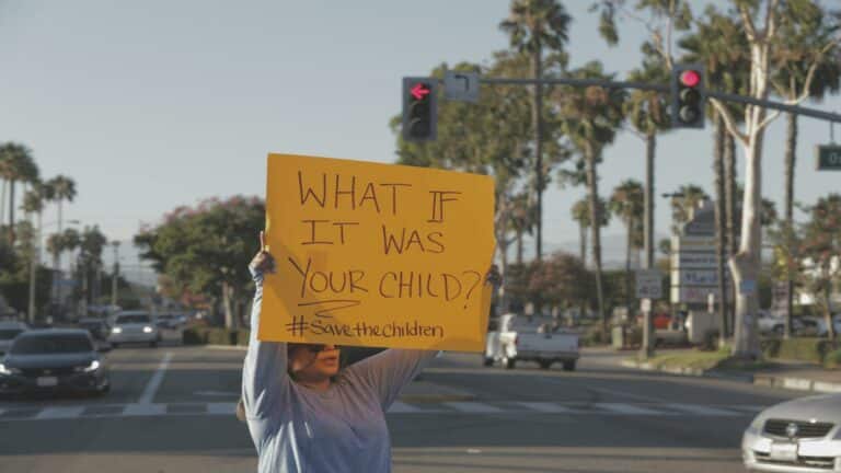 Person holding yellow protest sign with text 'What if it was your child? #SaveTheChildren' on palm tree-lined street