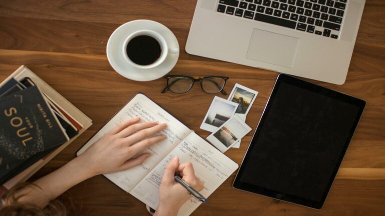 Person writing in journal with laptop, coffee, tablet and polaroid photos on wooden desk