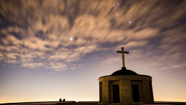 Small stone chapel with cross silhouetted against starry night sky with dramatic clouds, two people sitting in silhouette