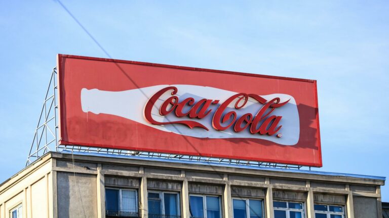 A Coca-Cola billboard mounted on top of a building with a red background and white logo.