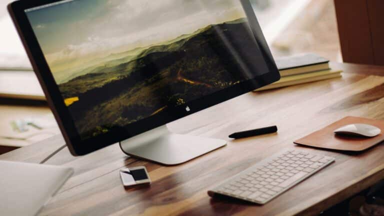 Professional desktop workspace with Apple monitor displaying mountain landscape, wireless keyboard, mouse, and stylus on wooden desk