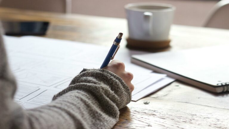 Person in grey sweater writing on paper with coffee cup in background