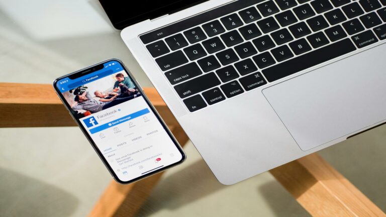 iPhone displaying Facebook app interface next to MacBook keyboard on wooden surface