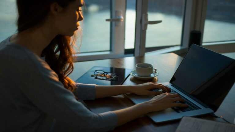 A person working on a laptop with a cup of coffee beside them at a desk near a window.