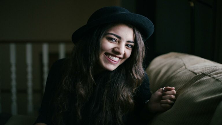 A woman with long hair wearing a black hat, smiling warmly while sitting on a couch.