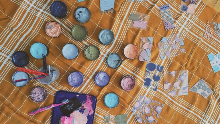 Various pastel-colored paint containers arranged on an orange and white checkered fabric with painting tools