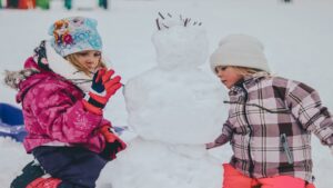 Two children dressed in winter clothing building a snowman outdoors, representing playful winter activities.