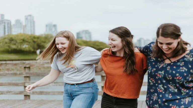 Three women walking arm in arm on a wooden pier with a cityscape in the background, representing teamwork and creativity in web design London.