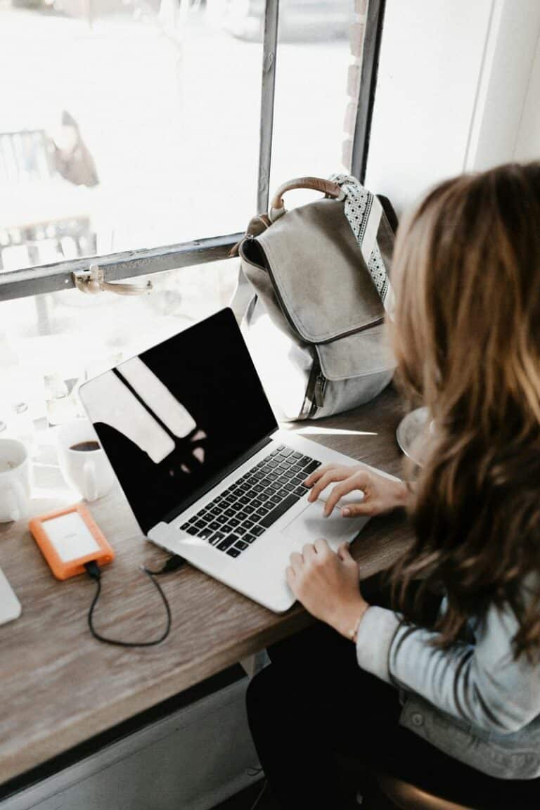 Find top-rated website design near me for your business. Woman working on a laptop with a coffee cup and a backpack nearby.